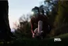 A woman is tying her shoe while wearing a headlamp in a natural outdoor setting at dusk. Náhled obrázku produktu