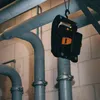 A black and orange LED work light is hanging from a hook, suspended from pipes in what appears to be a basement or utility area. Produktbild-Vorschau