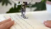 A close-up shot of a sewing machine needle stitching a wavy line on a piece of fabric. A person's hands are visible guiding the fabric. Produktbild-Vorschau