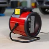 A red electric heater with a black stand is placed on a concrete floor in what appears to be a garage or workshop, with parts of cars visible in the background. Náhled obrázku produktu
