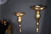 Two ornate gold wall consoles are mounted on a dark gray wall. The left console holds a pair of rings, and the right console holds a small dish with some decorative items. A partially visible clock is in the upper left corner, and a black table with gold t Produktbild-Vorschau