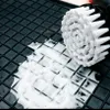 A close-up of a scrubbing brush with white bristles on a black textured surface with some white foam. Produktbild-Vorschau
