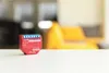 A small red electronic device with blue terminals sits on a white surface, with a blurred yellow chair in the background. Produktbild-Vorschau