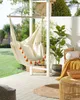A hammock chair with orange pom-poms hangs on a patio next to a potted plant and a wicker table. Produktbild-Vorschau