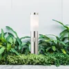 A modern stainless steel outdoor lamp with a frosted glass shade stands in a garden bed. Voir les images du produit