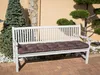 A white garden bench with a brown cushion sits on a brick patio, next to a tall evergreen tree and a decorative bonsai tree. Náhled obrázku produktu