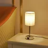 A modern LED nightstand lamp with a linen shade and USB charging ports is placed on a white nightstand next to a bed. Produktbild-Vorschau