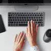 A top-down view of a person's hands typing on a wireless keyboard, with a smartphone and mouse visible. Náhled obrázku produktu