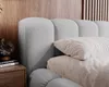 A close-up view of a grey upholstered bed with pillows and a bedside table with books. Náhled obrázku produktu