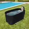 A woman is relaxing in a black portable bathtub filled with water, placed on a grassy lawn next to a swimming pool. Náhled obrázku produktu