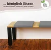 A close-up view of a wooden bench with gray seat cushions, set against a white wall and wooden floor. Produktbild-Vorschau