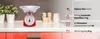 A red kitchen scale with a silver bowl sitting on a kitchen counter with plants and jars in the background. Náhľad obrázku produktu