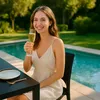 A woman is sitting at a table by a pool, holding a glass of champagne. Voir les images du produit