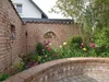 A garden scene with a brick wall, flowers, and a decorative iron window. Produktbild-Vorschau