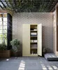 A tall beige cabinet with its doors open, showing neatly folded items and boxes inside, is placed on a tiled floor. Plants are visible to the left and right of the cabinet. The ceiling has hanging greenery. Anteprima dell’immagine prodotto