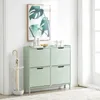 A mint green shoe cabinet with four flip-down drawers stands against a white wall, next to a white umbrella stand. A framed botanical print hangs above the cabinet. Produktbild-Vorschau