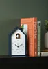A dark blue cuckoo clock sits on a black shelf next to two books, with a dark green wall in the background. Anteprima dell’immagine prodotto