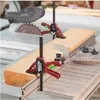 Two red and black Piher Einhandzwinge T-Track clamps are shown securing a wooden plank to a table saw. Podgląd zdjęcia produktu