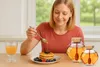 A woman is pouring honey from a spoon onto a stack of pancakes, with two honey jars and a glass of orange juice on the table. Náhled obrázku produktu