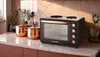 A black and gold countertop oven with two burners on top, placed on a kitchen counter with copper pots and a vase with greenery. Produktbild-Vorschau