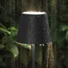 A black outdoor lamp is shown in the rain, with water droplets on its surface and water flowing from its base. Produktbild-Vorschau