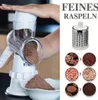 Weiße Drehreibe in Gebrauch, schreddert Schokolade in eine weiße Schüssel, mit einem separaten Bild der Feinschreddertrommel und Beispielen für geschredderte Schokolade, Erdnüsse und Kekse Voir les images du produit