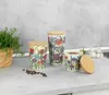 Three decorative storage canisters with bamboo lids are arranged on a kitchen countertop next to a potted herb and scattered coffee beans. Produktbild-Vorschau