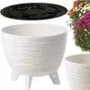 A white planter with a textured exterior and three legs, shown with a black insert and a cluster of colorful flowers in the background. Another planter of the same style is partially visible in the foreground. Produktbild-Vorschau