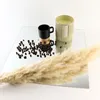 A still life arrangement on a mirrored surface, featuring coffee beans, a black mug with a gold accent, a candle, and a dried pampas grass plume. Produktbild-Vorschau