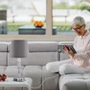 A woman is sitting on a couch and using a tablet, with a table lamp on a side table next to her. Produktbild-Vorschau