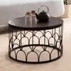 A round black coffee table with a decorative metal base, placed on a light-colored rug next to a sofa. Two glasses and a teapot are on the table. Produktbild-Vorschau