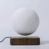 A levitating moon lamp with a textured white surface is shown floating above a wooden base. A black power cord is visible extending from the base. Produktbild-Vorschau
