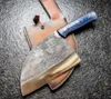 A cleaver knife with a blue and black patterned handle rests on a brown leather sheath, set against a dark, textured surface. Anteprima dell’immagine prodotto