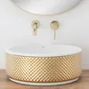 A close-up shot of a decorative washbasin with gold accents, mounted on a wooden surface, with gold faucets and a mirror in the background. Náhled obrázku produktu