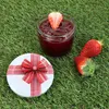A jar of strawberry jam with a strawberry slice on top, two strawberries, and a gift-wrapped lid on a grassy surface. Produktbild-Vorschau