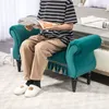 A person is sitting on a teal upholstered bench, reading a magazine. The bench has decorative tassels along the bottom and is placed in a room with a bed in the background. Náhľad obrázku produktu