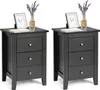 Two identical black nightstands are shown against a white background. Each nightstand has three drawers with gold knobs. On top of each nightstand, there is a framed picture, a small vase with dried flowers, and a book. Podgląd zdjęcia produktu
