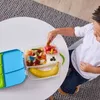 A young boy is sitting at a table, reaching into a lunchbox filled with food. The lunchbox is open, showing compartments with fruit, yogurt, and other food items. A banana is placed in one of the compartments. Náhľad obrázku produktu