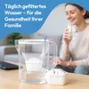A woman drinks filtered water from a glass next to a water filter pitcher. Produktbild-Vorschau