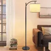 A floor lamp is placed next to a sofa and a stack of books, with a window in the background. Produktbild-Vorschau