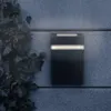 A black rectangular wall lamp with two illuminated strips is mounted on a textured gray wall, with ivy growing on the left side. Produktbild-Vorschau