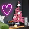 A pink Christmas tree with white star ornaments and wooden decorations, next to a neon pink heart light, two glasses of champagne, and a decorative ball on a wooden board. Produktbild-Vorschau