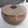 A round, hammered copper coffee table with two small cups and a coffee pot on top, placed on a light wooden floor.