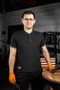 A man wearing a black polo shirt and orange gloves stands in a workshop, with tools and equipment in the background. Produktbild-Vorschau