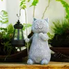 A garden statue of a cat holding a lantern, surrounded by plants. Produktbild-Vorschau