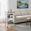A multi-tiered storage cart with a wooden top is shown in a living room setting next to a sofa. The cart contains various items like books, a plant, and folded towels. A painting hangs on the wall above the sofa. Voir les images du produit