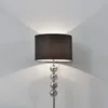 A floor lamp with a black shade and a silver base made of stacked glass balls, standing against a light gray wall. Produktbild-Vorschau