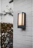 An outdoor wall light with a motion sensor is mounted on a white brick wall, with a blurred bush in the background. Anteprima dell’immagine prodotto
