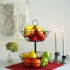 A two-tiered metal fruit basket filled with various fruits, placed on a table with decorative towels and candles in the background. Produktbild-Vorschau