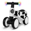 A white and black cow-patterned balance bike with colorful lights on the handlebars, shown against a white background. Produktbild-Vorschau